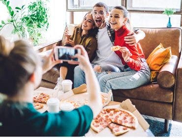 Cheerful smiling people embracing and sitting on couch by table with delivered pizza and cup while woman taking photo on smartphone in living room